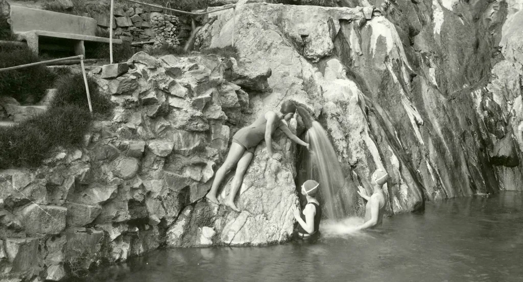 Three women are surrounding a small waterfall that is flowing into the spring. Two are standing in the water and one is leaning on the rock wall.