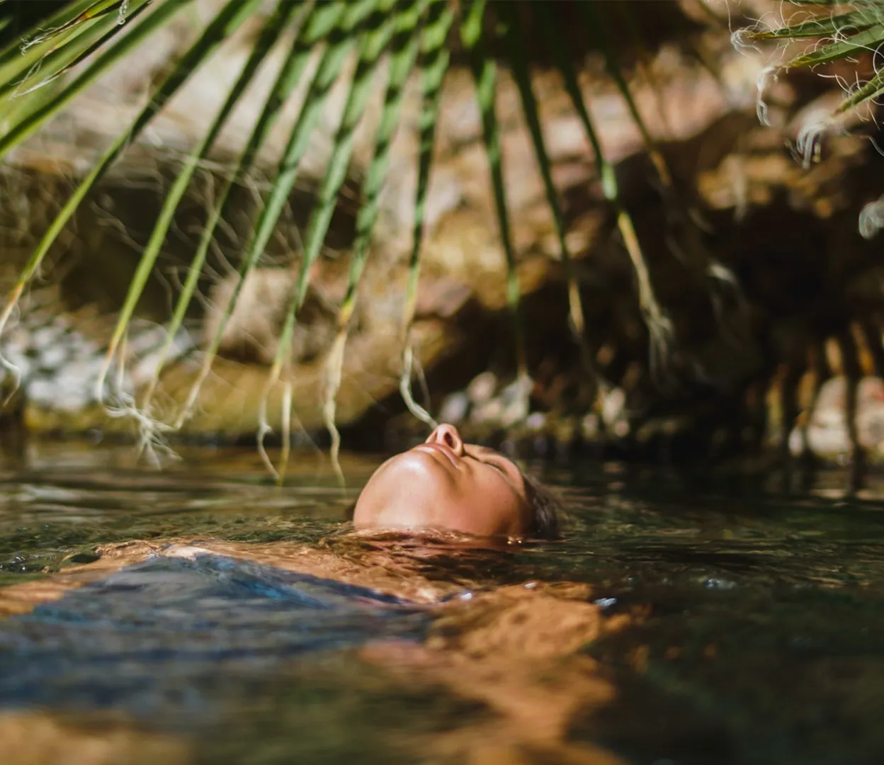 A closeup image of a woman floating in a hot spring with a palm close to her face.