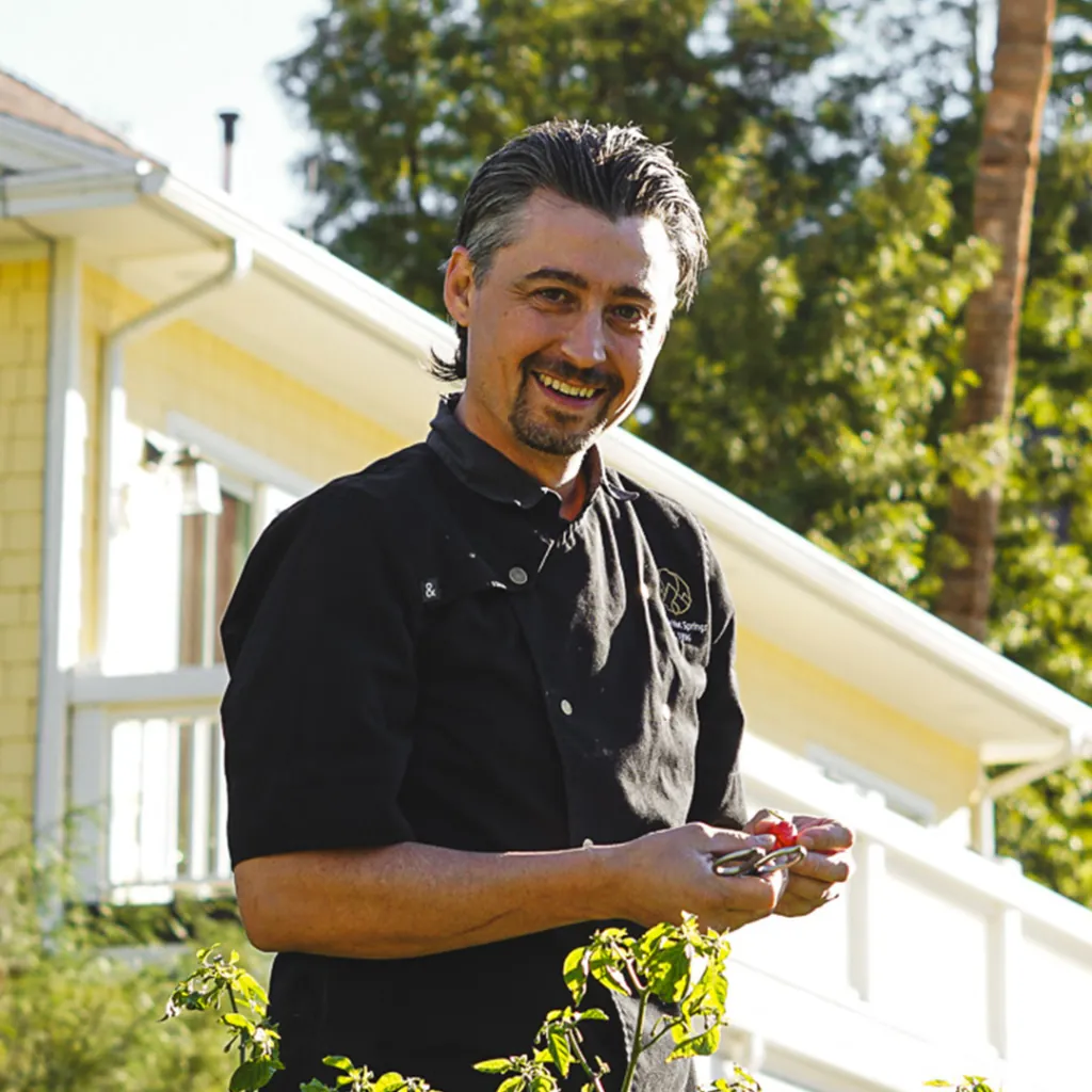 Chef Chris Knouse holds a red tomato outdoors near a yellow house and greenery.
