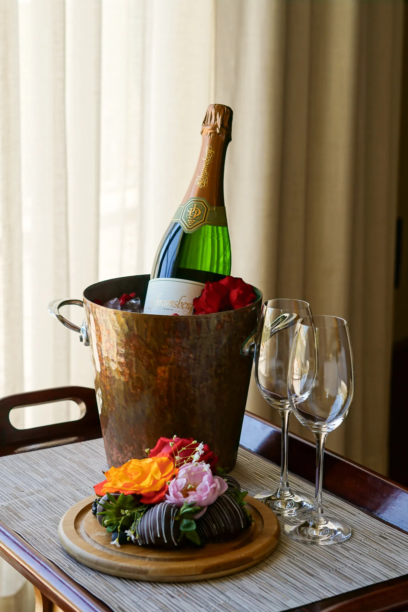 Champagne in an ice bucket with rose petals, two wine glasses, and chocolate-covered strawberries.