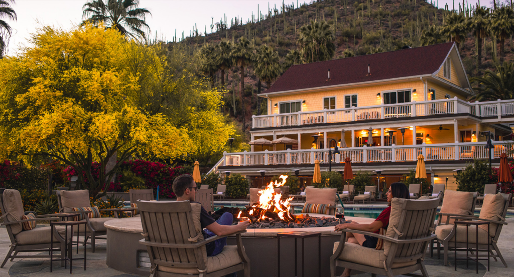 Two people sitting around a firepit with a yellow resort building in the background.