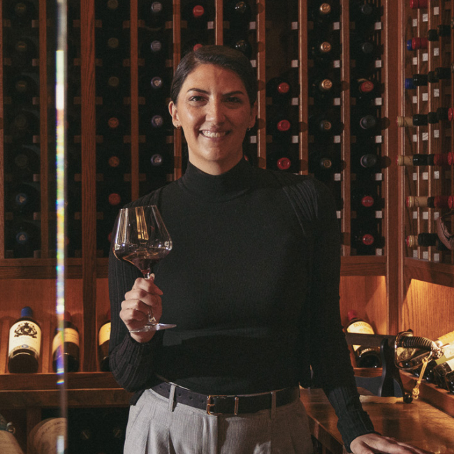 Sarah Foote holding a glass of red wine in a warmly lit wine cellar.