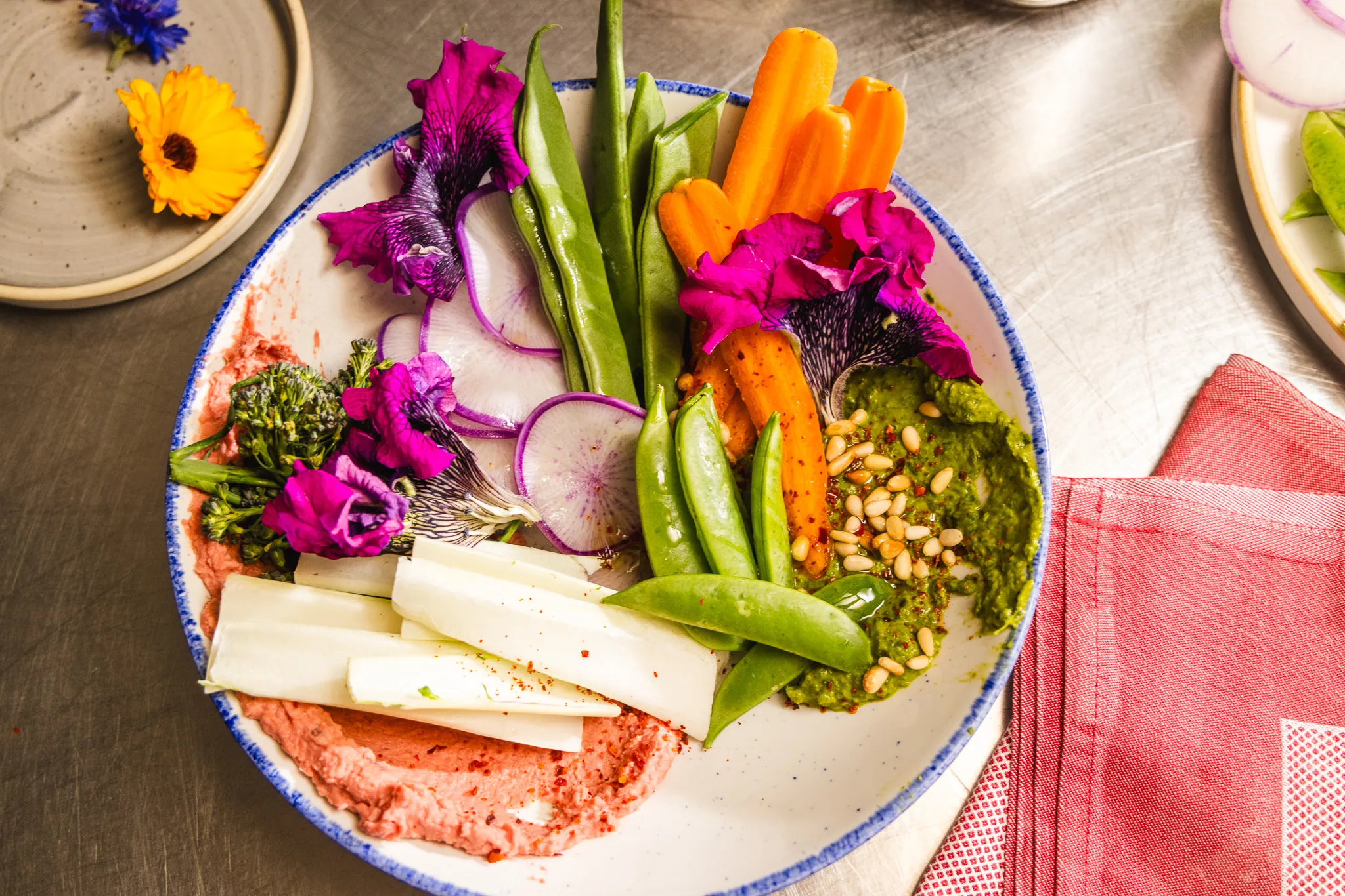Colorful plate with fresh veggies, hummus, pesto, edible flowers, and pine nuts on a rustic table.