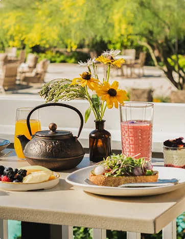 A cozy breakfast table at Harvest with a teapot, fresh food, flowers, and drinks in a sunny garden.
