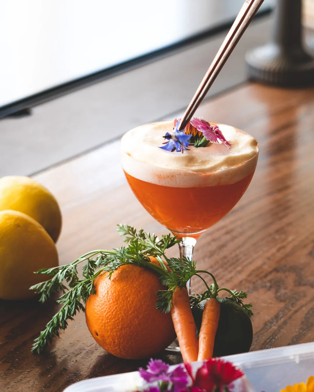 An orange cocktail with edible flowers, served with carrots and an orange on a wooden table.