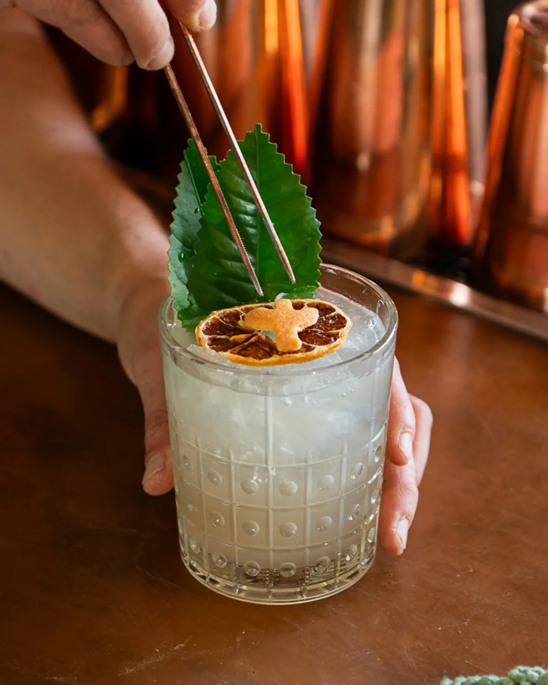 A cocktail with a dried orange slice and fresh leaf garnish, held in a textured glass.